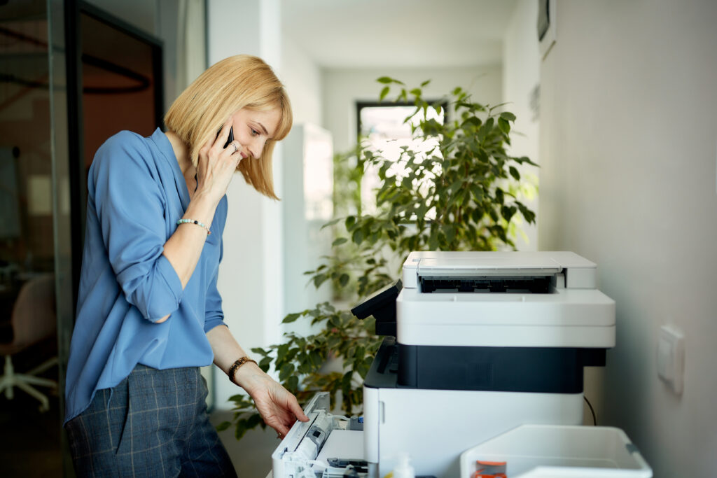 Smiling businesswoman printing documents while talking mobile phone in the office.